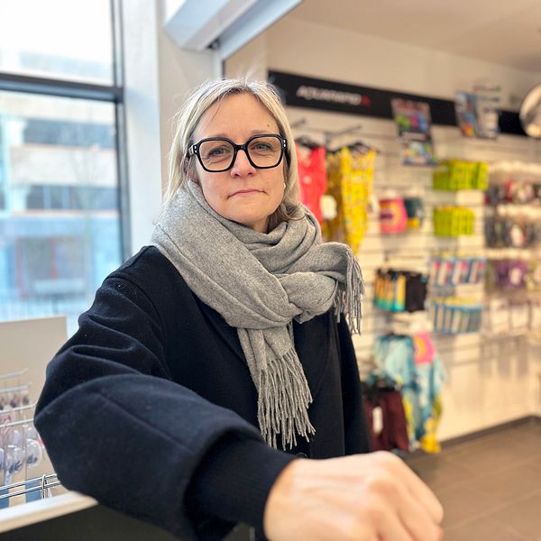 Badhuschef Åsa Säfsten Boman från Umeå kommun i vinterkläder står vid en butikshylla med simdräkter och andra produkter på bakre väggen.