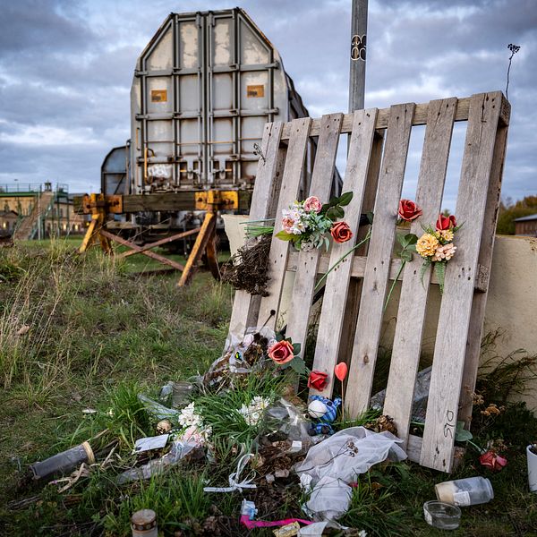 Minnesplats vid järnväg med blommor och ljus, gammalt tåg i bakgrunden.
