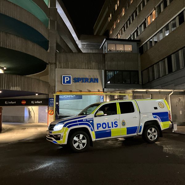 Polisbil parkerad vid ingången till ett parkeringshus med skylten ”SPIRAN” synlig i bakgrunden, omgiven av moderna byggnader i nattbelysning.