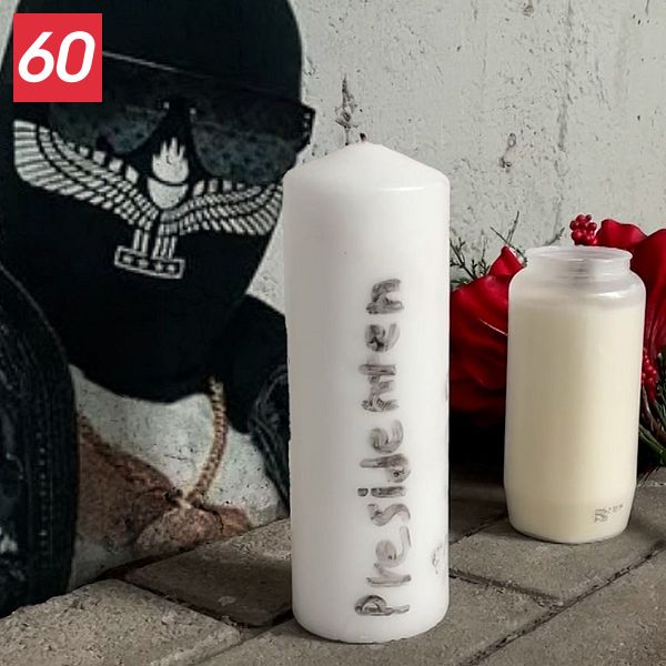 En man med en svart mask och solglasögon är klistrad mot en vägg. Framför honom finns två vita ljus och en röd ros placerade på marken. Denna scen kan tolkas som en hyllning eller minnesstund för en avliden person.