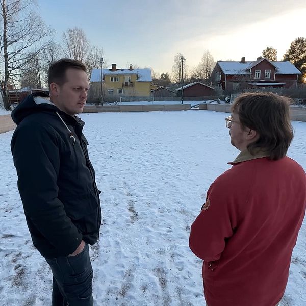 Två personer står i en snötäckt miljö med en byggnad i bakgrunden. Den ena personen är klädd i en svart jacka och den andra i en röd tröja. Trädkantade områden omger platsen och det är en klar himmel bakom dem.