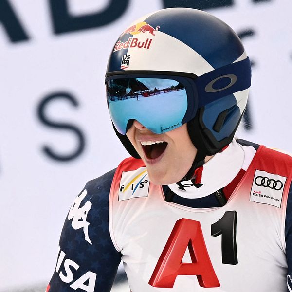 En alpin skidåkare i tävlingsdräkt med sponsormärken, förbereder sig för en nedfart. Skidåkaren har på sig en hjälm och en röd nummerlapp med bokstaven ”A1”. I bakgrunden syns en del av en stor reklamskylt med text.