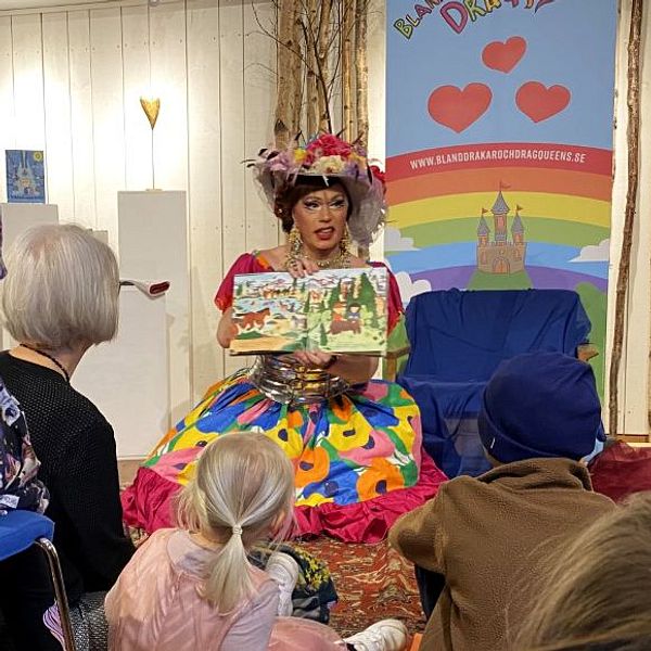 Person klädd i färggrann klänning läser bok för barn på bibliotek. Barnen sitter på golvet.
