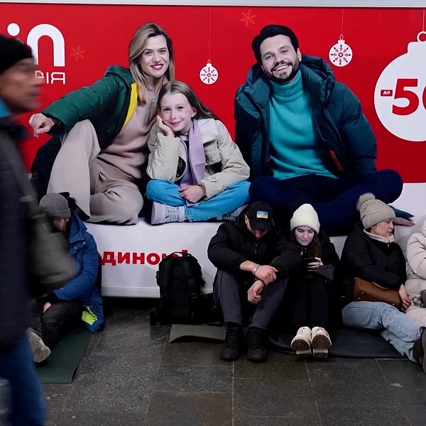 En grupp personer sitter på golvet i en tunnelbanestation, med en stor julskylt i bakgrunden som annonserar rea. Skylten är dekorerad med snöflingor och julgranartade motiv. Människorna är klädda i vinterkläder och några använder mobiler.