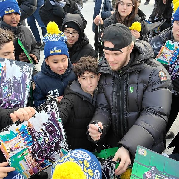 En grupp unga människor samlade runt en person som signerar autografer. De håller färgglada affischer och många av dem bär mössor och jackor som skydd mot kylan.