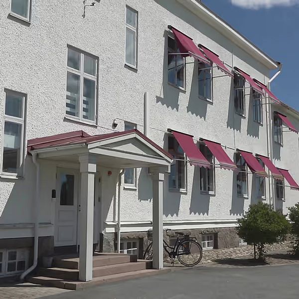 En vit byggnad med rödbruna markiser och en entré med tak. Det finns fönster på fasaden och en cykel stående på en sidogång. Grönska och buskar omger byggnaden, och i bakgrunden syns en annan byggnad och en blå himmel med några moln.