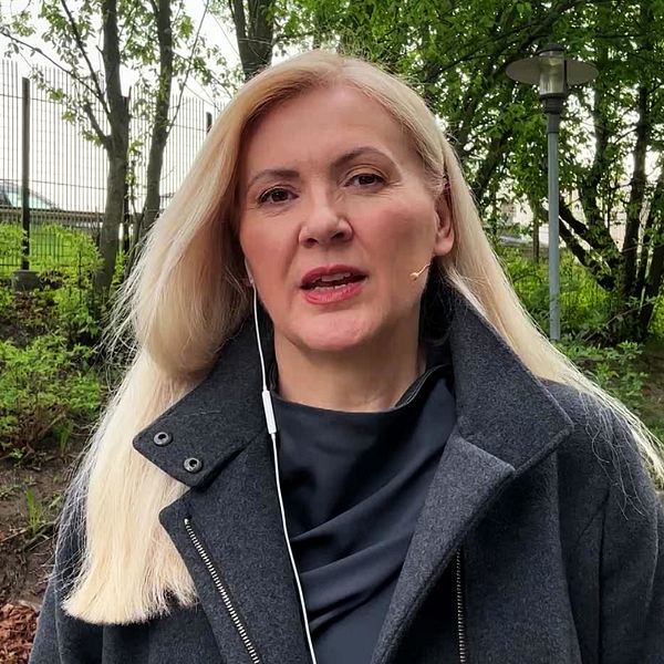 Kvinna med långt blont hår står utomhus på gångväg med gröna träd i bakgrunden.