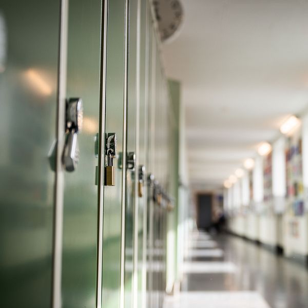 En lång korridor i en skola med gröna skåp på vänster sida, som är låsta med hänglås. Längs väggarna hänger färgglada affischer eller konstverk, och det finns mjukt ljus som strömmar in genom fönsterna.