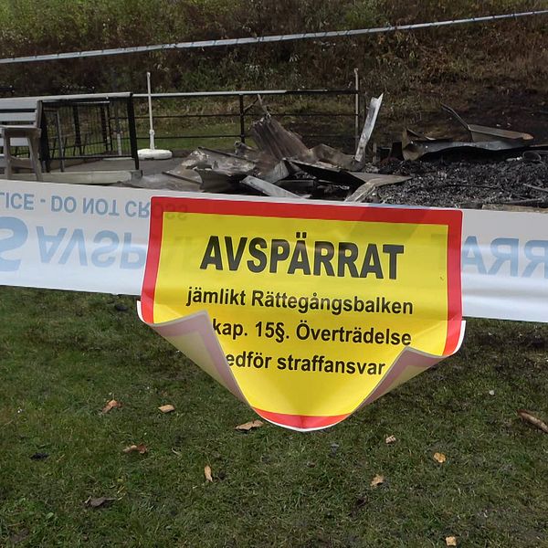 Avspärrningsband framför en brandskadad struktur på gräsmark.