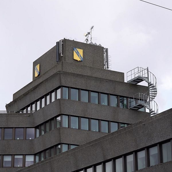 Grå kontorsbyggnad med spiraltrappa utvändigt och gula kommunlogotyper på toppen.