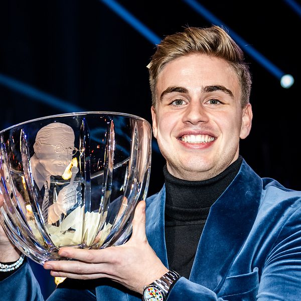 Personen i en blå sammetjacka håller upp en stor, klar glasbowl med en trofé inuti. Bakgrunden har starka ljusstrålar som skapar en festlig stämning.