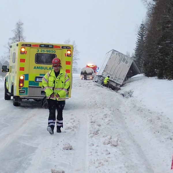 En lastbil ligger i diket intill en snöig väg. Framför står en ambulans och en man med gul väst kommer gåendes mot kameran.