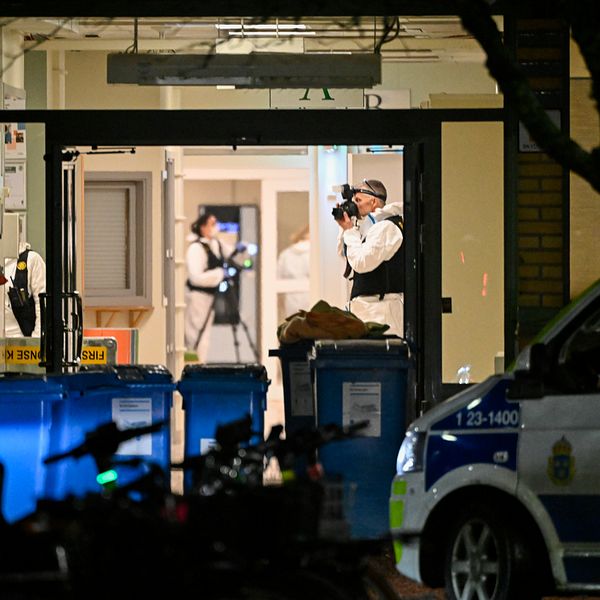 Kriminaltekniker arbetar inne i en byggnad, fotograferar och undersöker platsen. I förgrunden står en polisbil parkerad och flera soptunnor syns vid ingången.