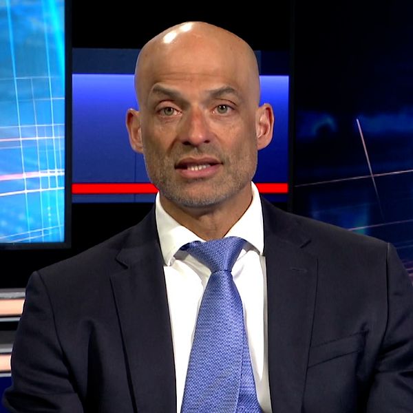 Person i kostym i tv-studio med blå bakgrund.