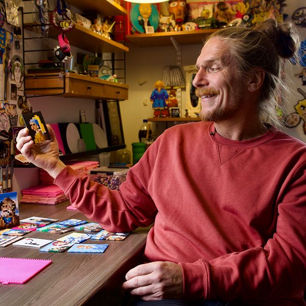 En person sitter vid ett bord i en kreativ arbetsmiljö, omgiven av färgglada dekorationer och konstverk på väggarna. På bordet ligger en uppsättning bilder och en rosa pärlplatta. Personen håller upp en av bilderna.