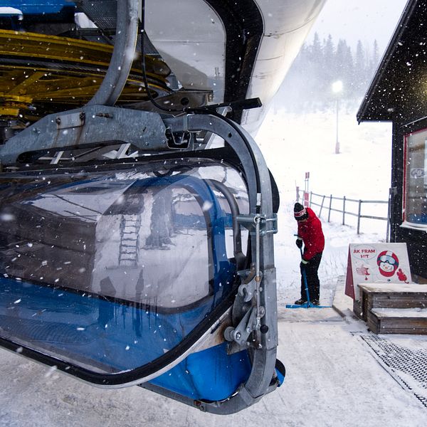 En liftkorg hänger från en skidlift i snöfall i Duved, medan en liftvärd sopar av påstigningsytan. I bakgrunden syns en skidbacke.
