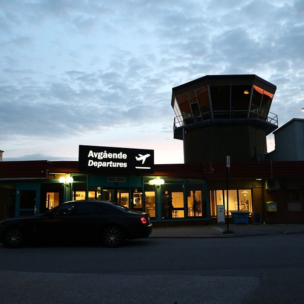 Bil utanför flygplatsens avgångshall i kvällsljus med upplyst skylt och flygledningstorn.
