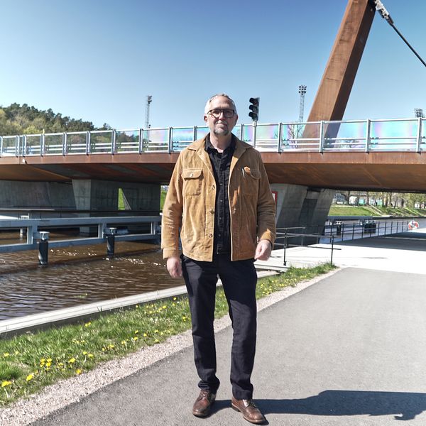 Man i brun jacka står på gångväg framför träbro vid kanal en solig dag.
