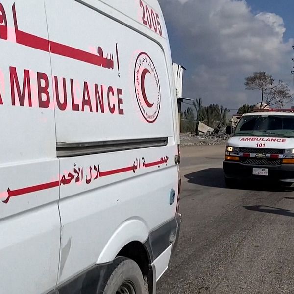Två ambulanser står på en väg med tydliga röda kors och arabiska bokstäver på sidorna. Två personer i uniformer går bredvid ambulanserna under en molnig himmel.