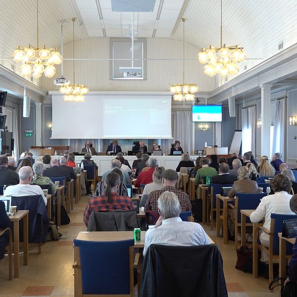 Möte pågår i stor konferenssal, deltagarna sitter vid bord och lyssnar på talare framme.