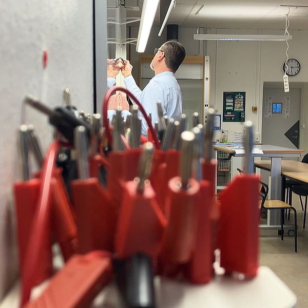 Ett klassrum fullt med elever och föreläsningsmaterial. I förgrunden syns kablar och elektronikverktyg, medan en man i bakgrunden skriver på en whiteboard.