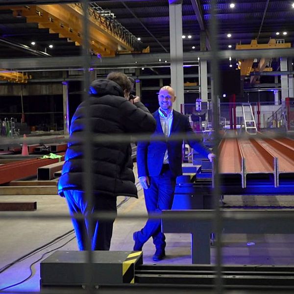En man i kostym står och pratar med en annan person iförd en vinterjacka i en stor industrilokal. I bakgrunden syns stålramar och kranar, samt belysning som skapar en dramatisk atmosfär. Bilden är tagen genom ett galler.