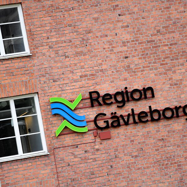 Logotypen för Region Gävleborg på en tegelvägg, med en grafisk design som inkluderar blå och gröna färger samt texten ”Region Gävleborg”. Flera fönster är synliga i närheten.