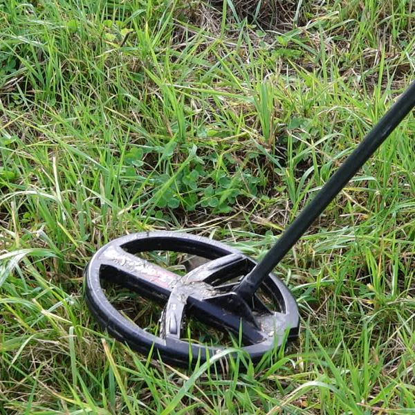 En metalldetektor som står i gräset, med fokus på den rundade sökspolen nära markytan. Gräset är frodigt och grönt runt detektorn.