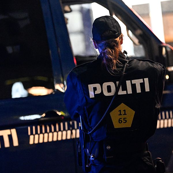 Polis i uniform står med ryggen mot kameran vid en blå polisiär bil, med ordet ”POLITI” synligt på både bilen och uniformen.