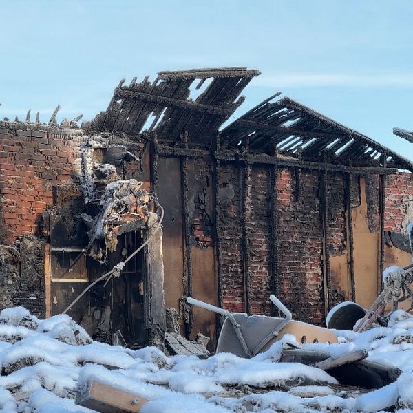 Resterna av en branddrabbad byggnad är synliga, där tegelväggar är blottade och taket har kollapsat. Snö täcker marken runtomkring, vilket tyder på kallt väder. Det finns skräp och rivningsmaterial spritt på marken, vilket visar att byggnaden har genomgått betydande skador.