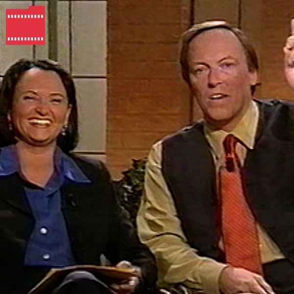 Två personer sitter i en studio, en av dem håller upp ett finger och verkar tala.