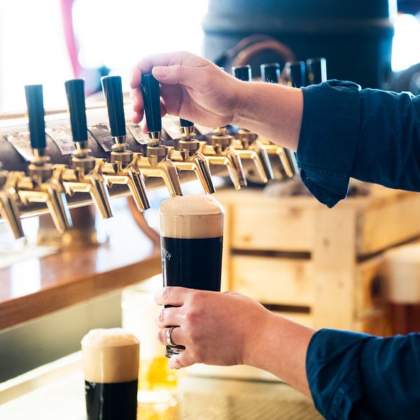 En person häller upp ett mörkt öl från en tappkran i en bar, med flera tappkranar synliga i bakgrunden. Ölet har ett stort, skummande huvud och det finns flera glas med öl på bardisken.