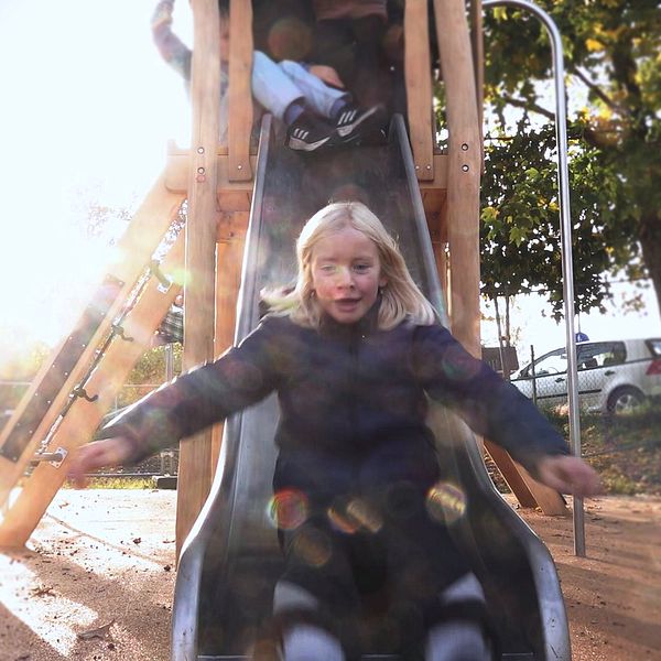 Ett barn åker ner för en rutschkana i en lekpark, omgiven av andra barn som leker. Solens ljus skiner starkt.