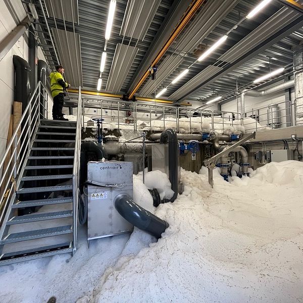 En person med skyddsutrustning står på en trappa bredvid stora mängder skum på golvet i en industriell anläggning.