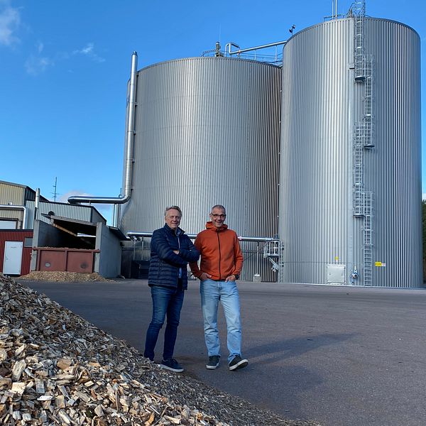 Två personer står framför en industriell anläggning med stora cylindriska tankar i metall. I förgrunden syns en hög med träflis. Bakgrunden består av en klar blå himmel och skog.