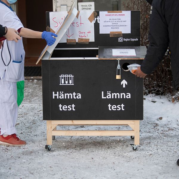 Personer står vid en utomhusstation för hantering av covid-19-tester. En skylt visar två sektioner, ”Hämta test” och ”Lämna test”, där en person i sjukvårdskläder tar hand om testerna. Marken är täckt av snö.
