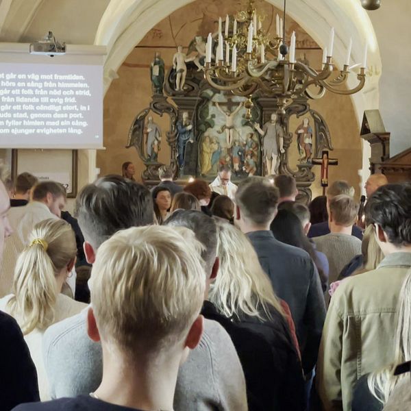 Människor samlas i en kyrka för en ceremoni, med en stor altarvägg och religiösa konstverk i bakgrunden. På en skärm i förgrunden visas text, vilket tyder på en presentation eller hymn. Atmosfären är stillsam och reflekterande.