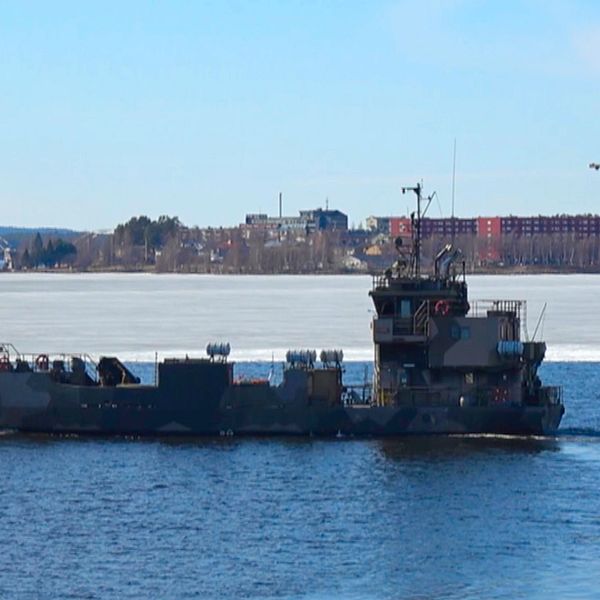 Militärt fartyg kör på vattnet med stad och is i bakgrunden.