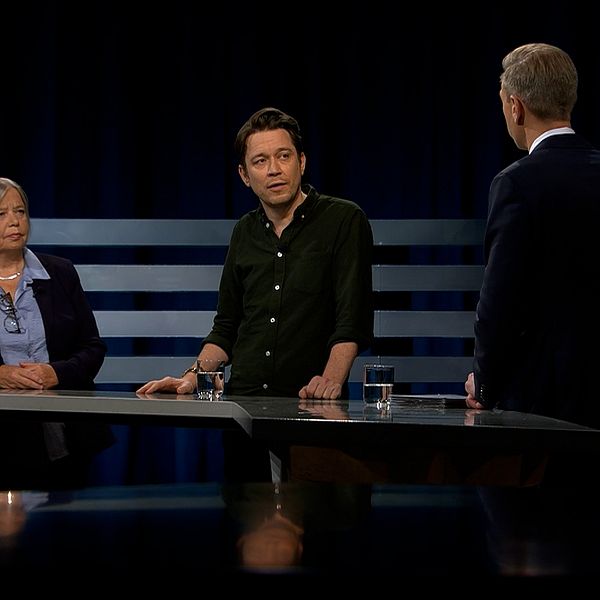Tre personer står vid ett podium under en studiodiskussion. Det finns glas vatten på bordet och en mörk bakgrund som framhäver scenen.