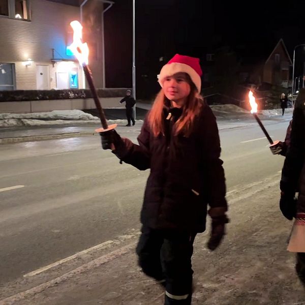 Två personer går längs en gata under natten, bärande på lågor som lyser upp omgivningen. De har på sig tomteluvor och är klädda i varma kläder. Vinterlandskapet har en del snö på marken och byggnader syns i bakgrunden.