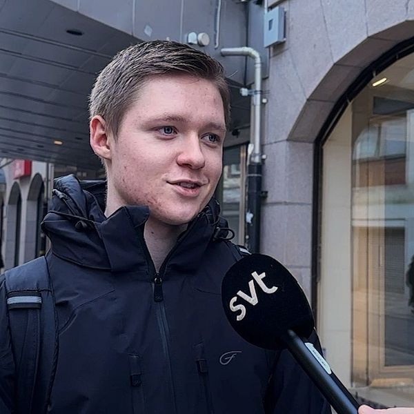En person står på en trottoar i en stadsmiljö, klädd i en mörk jacka och bär en ryggsäck. En mikrofon med SVT:s logotyp sträcks fram mot personen, som antagligen deltar i en intervju. I bakgrunden syns fler människor, byggnader och en parkerad bil.