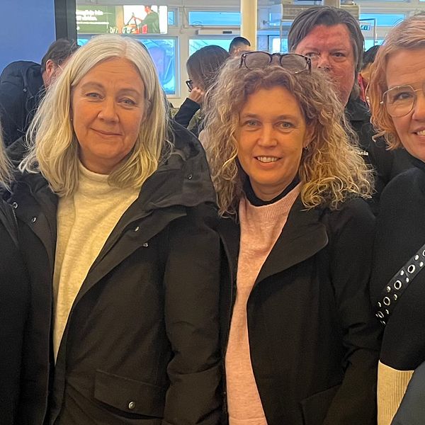 En grupp kvinnor står tillsammans i en livlig miljö, klädda i svarta och grå jackor. De ser ut att vara engagerade i en konversation och leende mot kameran. Bakgrunden visar en ljus plats med otydliga silhuetter av andra människor.