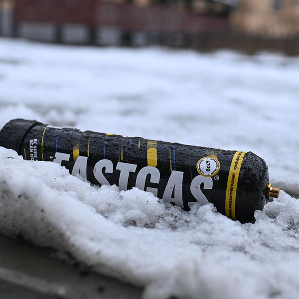 En svart flaska med texten ”FASTGAS” ligger på marken omgiven av snö i en urban miljö med byggnader i bakgrunden.