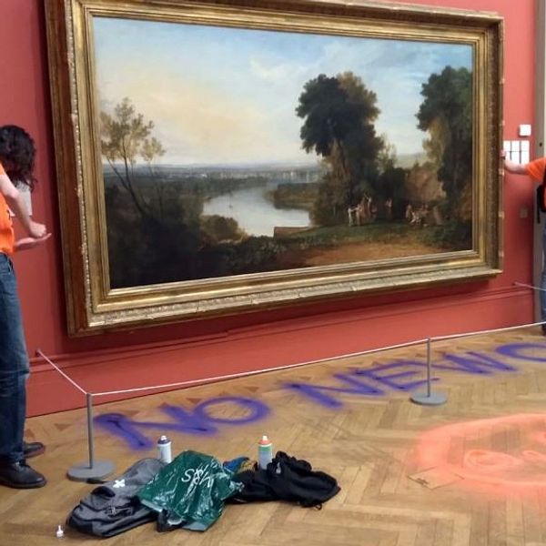 Två personer i orangea tröjor står framför en stor landskapsmålning på en vägg i ett konstgalleri. De verkar vara delaktiga i en protestaktion, där texten ”NO NEW OIL” är målad på golvet nedanför målningen. En av dem har en sprayburk i handen, medan deras väskor och utrustning ligger på golvet. Målningen föreställer en naturscen med träd och vatten.