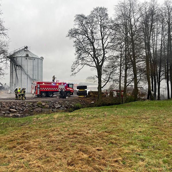 Brandmän arbetar vid en industribyggnad på landsbygden, omgiven av träd och gräs.