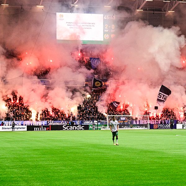 Fulsatt läktare fylld av bengalrök. Spelare ute på en fotbollsplan.