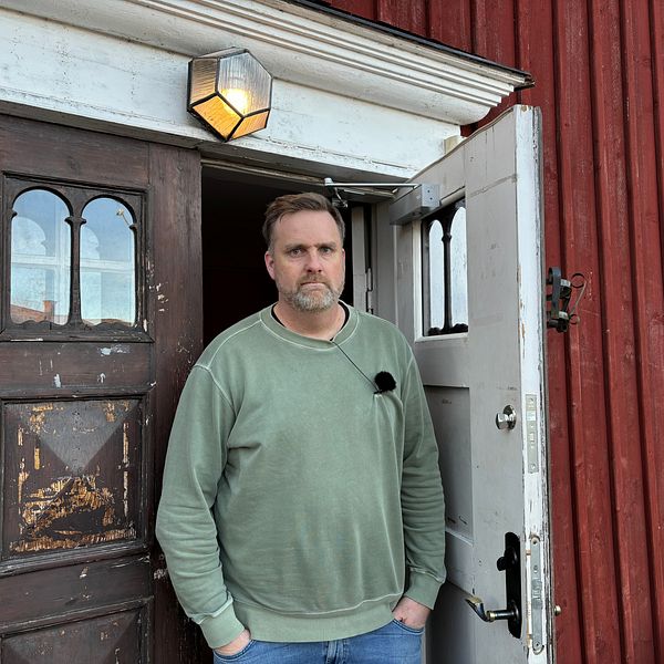 En man står i dörröppningen till en röd stuga. Dörren är öppen och på väggen ovanför dörren hänger en lampa. Bakom mannen syns en del av interiören i stugan.