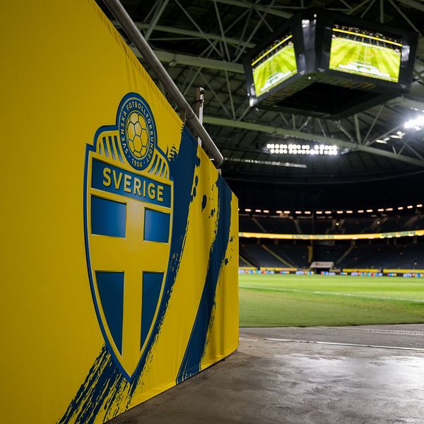 Insidan av Friends Arena i Stockholm med det svenska fotbollslandslagets emblem på en gul vägg i förgrunden och en nästan tom fotbollsplan upplyst i bakgrunden.