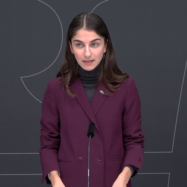 En person står vid ett podium under en presskonferens, iklädd en lila kappa och en mörk tröja. Det finns en mikrofon framför personen, och en mörk bakgrund med grafiska inslag syns bakom.