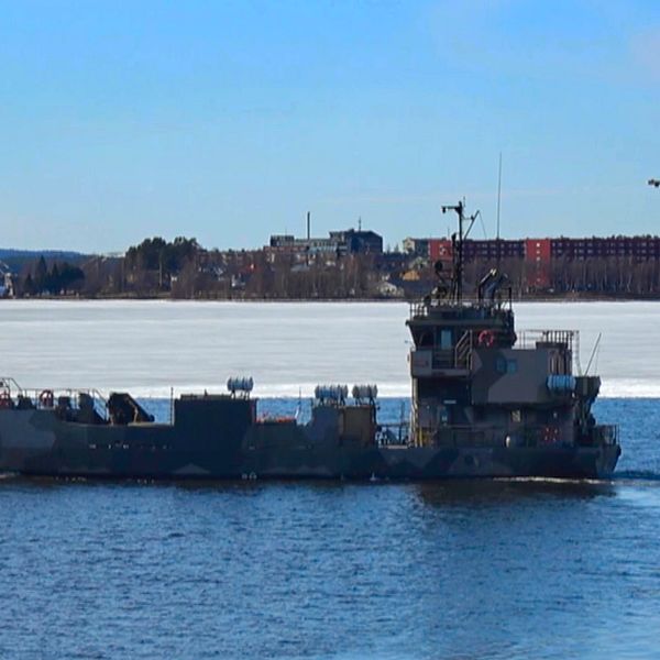 Militärbåt färdas på vattnet med is i bakgrunden och stadssilhuett längre bort.
